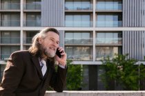 Вид збоку усміхненого бородатого зрілого чоловіка з сірим волоссям в стильному костюмі обговорюючи бізнес-задачі під час телефонної розмови, стоячи проти сучасних міських будівель — стокове фото