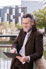 Positive respectable middle aged bearded man in formal outfit browsing tablet while standing on embankment in city — Stock Photo