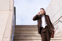 From below of confident mature male executive manager in classy suit with bag drinking takeaway coffee while walking down stairs in city — Stock Photo