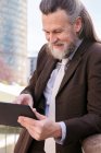 Позитивный респектабельный бородатый мужчина средних лет в формальной одежде просматривает планшет, стоя на набережной в городе — стоковое фото