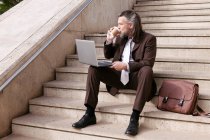 Middle aged bearded male entrepreneur in formal clothes sitting on stairway drinking takeaway coffee and working online on laptop in city — Stock Photo