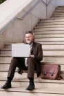 Positive middle aged bearded male entrepreneur in formal clothes sitting on stairway and working online on laptop in city — Stock Photo