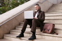 Positive middle aged bearded male entrepreneur in formal clothes sitting on stairway and working online on laptop in city — Stock Photo
