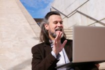 Знизу позитивного літнього бородатого чоловіка в елегантному костюмі, сидячи на сходах і махаючи рукою і кажучи привіт, маючи онлайн відеозустріч через ноутбук — стокове фото