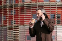 Uomo barbuto dai capelli grigi in abito formale con tazza di caffè da asporto in mano parlando sul cellulare mentre in piedi sulla strada urbana — Foto stock
