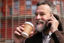 Hombre barbudo de pelo gris en traje formal con taza de café para llevar en la mano hablando por teléfono móvil mientras está de pie en la calle urbana - foto de stock