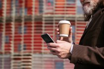 Вид сбоку на седого бородатого человека в костюме, пьющего кофе на вынос и просматривающего мобильный телефон на городской улице — стоковое фото