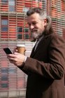 Side view of gray haired bearded man in formal suit drinking takeaway coffee and browsing mobile phone on urban street — Stock Photo