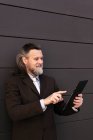 Елегантний бородатий зрілий чоловік у стильному формальному костюмі, що стоїть біля сірої стіни та переглядає Інтернет на цифровому планшеті — стокове фото