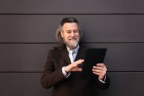 Элегантный бородатый зрелый мужчина в стильном костюме стоит рядом с серой стеной и просматривает интернет на цифровом планшете — стоковое фото