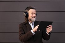 Содержание седовласый бородатый мужчина в формальном костюме и беспроводные наушники с помощью планшета во время общения в Интернете против серой стены — стоковое фото