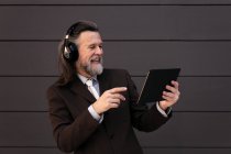 Contenido Hombre barbudo de pelo gris en traje formal y auriculares inalámbricos usando tableta mientras se comunica en línea contra la pared gris - foto de stock