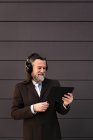 Content gray haired bearded male in formal suit and wireless headphones using tablet while communicating online against gray wall — Stock Photo