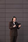 Content gray haired bearded male in formal suit and wireless headphones using tablet while communicating online against gray wall — Stock Photo