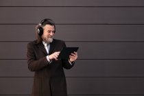Contenido Hombre barbudo de pelo gris en traje formal y auriculares inalámbricos usando tableta mientras se comunica en línea contra la pared gris - foto de stock