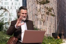 Side view of happy successful gray haired bearded male in elegant suit waving hand and saying hello while having online video meeting via laptop sitting on urban street — Stock Photo