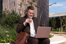 Щасливий успішний сіро-волохатий бородатий чоловік в елегантному костюмі виймає каву за допомогою ноутбука, сидячи на міській вулиці — стокове фото
