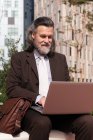 Feliz éxito de pelo gris barbudo macho en traje elegante usando el ordenador portátil mientras está sentado en la calle urbana - foto de stock