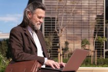 Vista lateral de feliz éxito de pelo gris barbudo macho en traje elegante utilizando el ordenador portátil mientras está sentado en la calle urbana - foto de stock