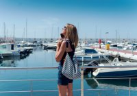 Vista lateral da fêmea encantada em pé no porto com veleiros e falando no telefone celular no verão no dia ensolarado — Fotografia de Stock