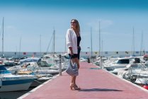 Happy female in summer outfit walking along promenade near sea with moored yachts — Stock Photo