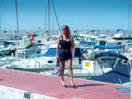 Hembra positiva caminando a lo largo del terraplén en el puerto con yates amarrados en un día soleado y disfrutando de las vacaciones de verano - foto de stock