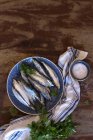 Vista superior de um prato com sardinhas frescas em uma mesa de madeira ao lado de salsa e uma toalha de cozinha — Fotografia de Stock
