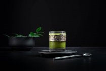 Stillleben-Komposition mit traditionellem orientalischem Matcha-Tee, serviert in Glasschale mit Metalldekor auf Tisch mit Keramikschalen und frischen grünen Blättern vor schwarzem Hintergrund — Stockfoto