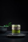 Still life composition with traditional oriental matcha tea served in glass cup with metal ornamental decor on table with ceramic bowls and fresh green leaves against black background — Stock Photo
