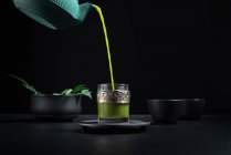 Chá matcha japonês saudável sendo derramado de bule verde em copo de vidro com decoração decorativa de metal durante a cerimônia de chá contra fundo preto — Fotografia de Stock