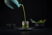 Thé matcha japonais sain versé de la théière verte dans une tasse en verre avec un décor ornemental en métal lors de la cérémonie du thé sur fond noir — Photo de stock
