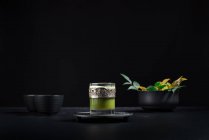 Stillleben-Komposition mit traditionellem orientalischem Matcha-Tee, serviert in Glasschale mit Metalldekor auf Tisch mit Keramikschalen und frischen grünen Blättern vor schwarzem Hintergrund — Stockfoto
