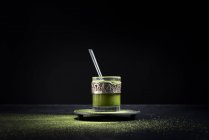 Thé matcha vert à base de plantes sain servi dans une tasse en verre avec décoration en métal sur une soucoupe saupoudrée de poudre sur une table noire — Photo de stock