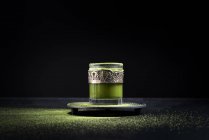 Té matcha verde herbal saludable servido en taza de vidrio con decoración de metal en platillo espolvoreado con polvo en mesa negra - foto de stock