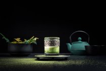 Gesunder grüner Kräutermatcha-Tee serviert in Glasschale mit Metalldekoration auf Untertasse bestreut mit Pulver auf schwarzem Tisch — Stockfoto