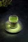 Dall'alto di sano tè verde matcha alle erbe servito in tazza di vetro con decorazione in metallo sul piattino cosparso di polvere sul tavolo nero — Foto stock