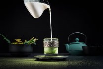 Frische Milch wird während der traditionellen Zeremonie aus dem Glas in eine Glasschale mit Matcha-Tee gegossen und auf den Tisch mit Teekanne und Geschirr gestellt — Stockfoto