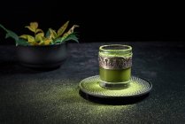 Зверху здоровий трав'яний зелений чай з мачами подається в скляній чашці з металевою прикрасою на блюдці, посипаної порошком на чорному столі — стокове фото