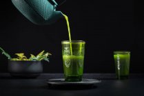 Gesunder japanischer Matcha-Tee, der während der Teezeremonie vor schwarzem Hintergrund aus einer grünen Teekanne ins Glas gegossen wird — Stockfoto