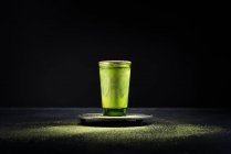 Té matcha verde herbal saludable servido en taza de vidrio con decoración de metal en platillo espolvoreado con polvo en mesa negra - foto de stock