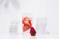 Agua sana y refrescante para desintoxicar con fresas frescas en rodajas maduras servidas en vidrio transparente contra la pared blanca con sombras - foto de stock