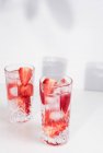Refrescante bebida de verano con fresas frescas en rodajas y cubitos de hielo con agua servida en vasos sobre mesa blanca - foto de stock
