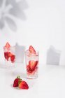 Освежающий летний напиток со свежей клубникой и кубиками льда с водой, подаваемой в стаканах на белом столе — стоковое фото