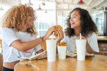 Amigos afroamericanos positivos sentados a la mesa con comida rápida y bebidas y disfrutando del fin de semana en la cafetería - foto de stock