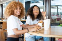 Позитивные афроамериканские подруги сидят за столом с фаст-фудом и напитками и наслаждаются выходными в кафе — стоковое фото