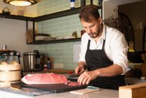 Сфокусований чоловік - кухар ріже сиру рибу за столом в азіатському ресторані і готує суші. — стокове фото