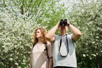 From below of male traveler near wife observing birds through binoculars in green woods in summer — Stock Photo
