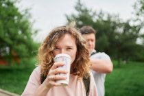 Charmante femelle aux cheveux bouclés profitant d'une boisson chaude dans une tasse en papier à emporter tout en regardant la caméra dans un parc d'été — Photo de stock
