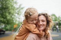 Милая маленькая девочка обнимает веселую мать в парке, проводя вместе выходные летом — стоковое фото