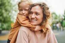 Милая маленькая девочка обнимает веселую мать в парке, проводя вместе выходные летом — стоковое фото